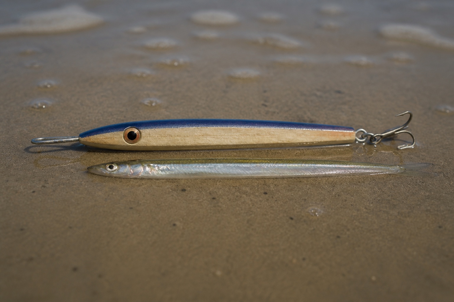 Slim wooden needlefish plug lying on wet sand beside a sand eel of similar length for size comparison